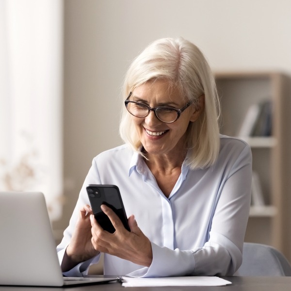Eine ältere Frau sitzt mit ihrem Smartphone vor ihrem Laptop und führt einen Videoanruf.